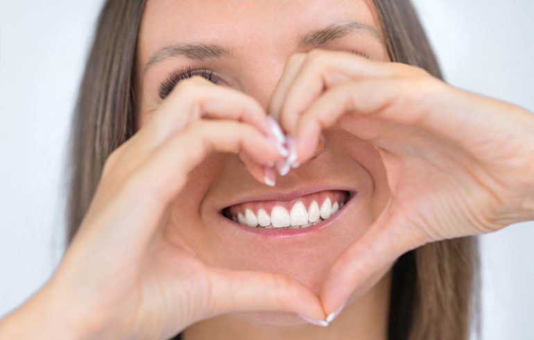 Broadly smiling woman with healthy teeth and gums holding hands put in shape of heart in front of her mouth.