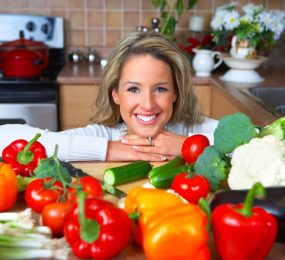 A woman with a perfect smile behind a kitchen counter covered with variety of vegetables.