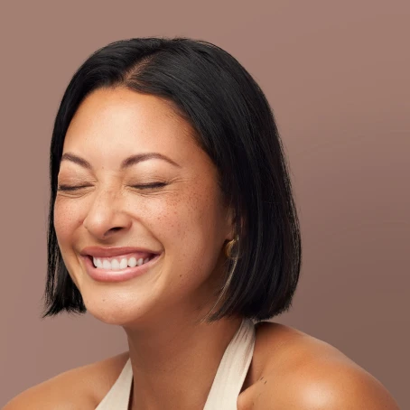 A person with short black hair smiling with closed eyes against a plain background.