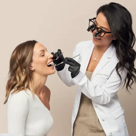 A dentist wearing magnifying glasses examines a patient's teeth using dental tools in an office setting.