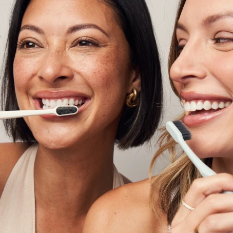 Two people smiling while brushing their teeth with black-bristled toothbrushes. They appear to be in a bright setting.