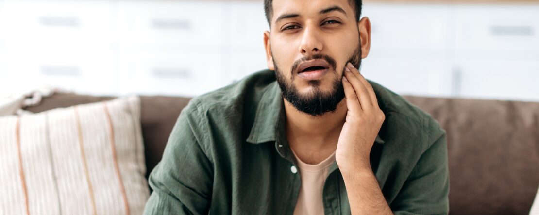 Frustrated unhealthy indian or arabian man in casual clothes, pressing cheek with hand, suffering toothache, caries, sits on a sofa in living room, needs the help of a dentist. Dental problems concept