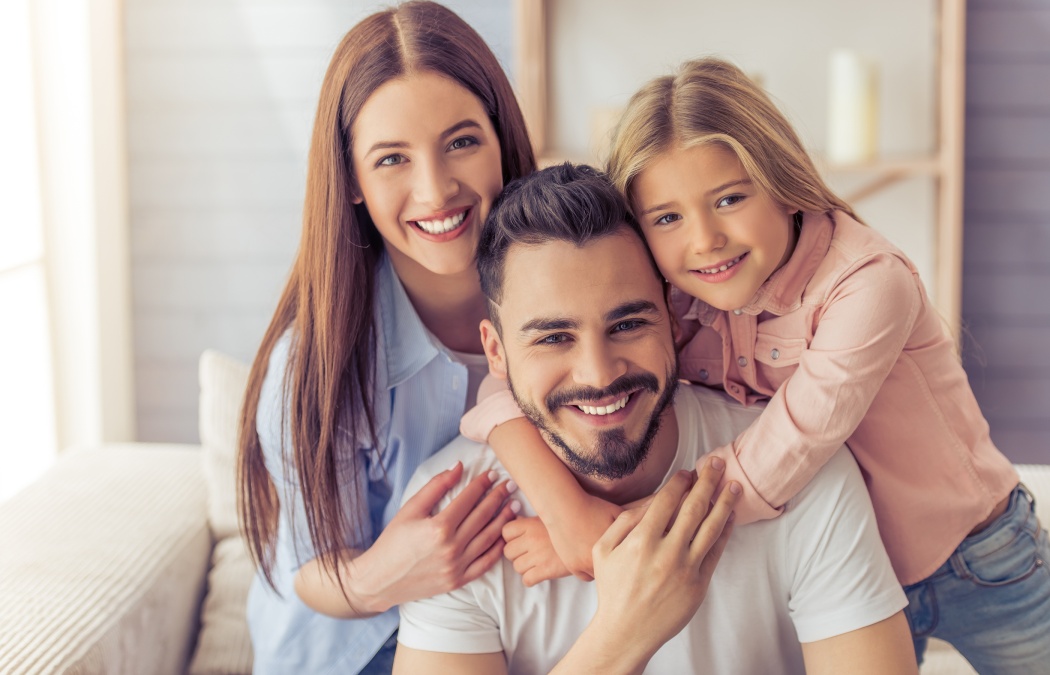 Portrait of beautiful young mother father and their daughter hugging looking at camera and smiling while sitting on sofa at home