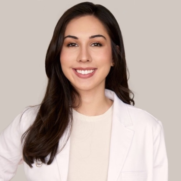 A woman with long brown hair, wearing a white lab coat and a light top, smiles at the camera against a plain beige background, reflecting the welcoming atmosphere of Les Belles NYC and their cosmetic dentistry expertise.