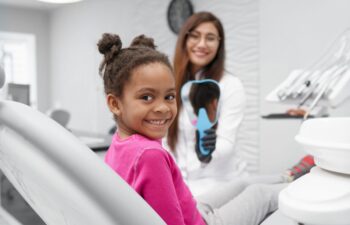 A dentist holding a mirror and a little smiling Afro-American girl sitting in a dental chair after the routine checkup and cleaning