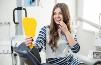 Beautiful young female patient with perfect white teeth sitting in dental chair looking in the mirror and checking up teeth after treatment at modern dental clinic. Dentistry and healthcare concept