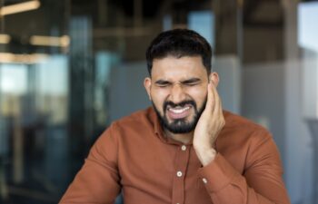 Young middle eastern indian man grimacing in pain, clutching his cheek from a severe toothache or jaw inflammation, urgent dental problem needing immediate care