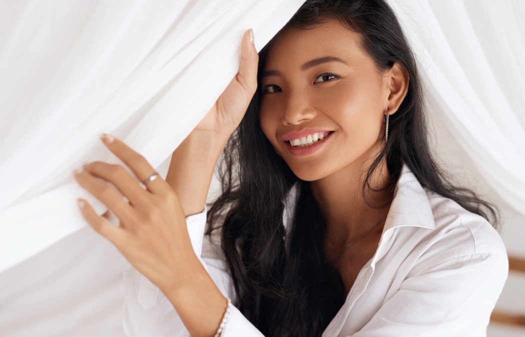 Happy Woman With Beautiful Face Smiling, Lying On Bed. Portrait Of Beautiful Young Female With Healthy Hair, Fresh Soft Facial Skin And Perfect White Smile On White Bedding. High Resolution.